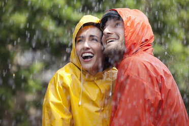 Glückliches Paar in Regenmänteln schaut in den Regen - CAIF19769