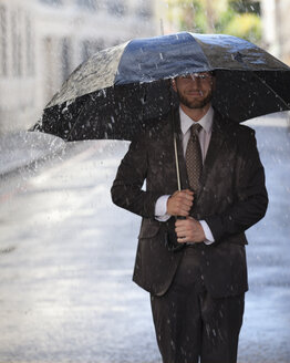 Glücklicher Geschäftsmann mit Regenschirm auf verregneter Straße - CAIF19777