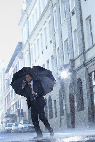 Geschäftsmann, der unter einem Regenschirm in einer regnerischen Straße mit seinem Handy telefoniert, lizenzfreies Stockfoto
