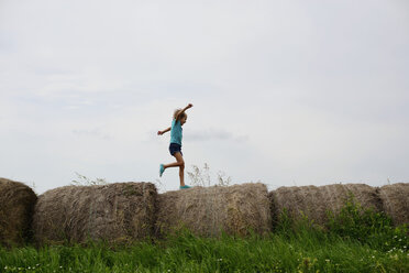 Verspieltes Mädchen läuft auf Heuballen auf einem grasbewachsenen Feld gegen den Himmel - CAVF10374