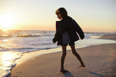 Mädchen in Jacke schaut auf das Meer, während sie am Strand bei Sonnenuntergang steht - CAVF10415