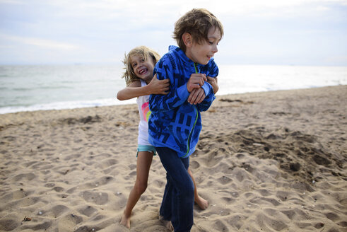 Verspielte Schwester kitzelt Bruder, während sie am Huntington Beach gegen den Himmel steht - CAVF10440