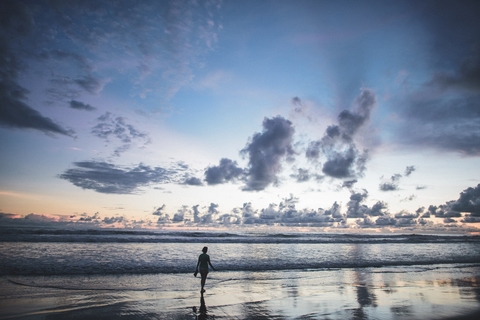 Rückansicht einer am Strand stehenden Frau gegen einen bewölkten Himmel bei Sonnenuntergang, lizenzfreies Stockfoto