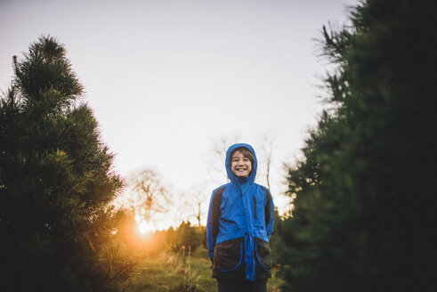 Glücklicher Junge mit Kapuzenjacke, der bei Sonnenuntergang auf einem Feld unter Kiefern steht - CAVF10479