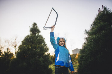 Portrait of girl holding hand saw while standing against pine trees on field against clear sky - CAVF10480