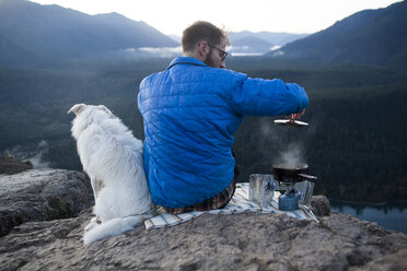 Rückansicht eines Mannes mit Hund, der auf einem Berg Essen zubereitet - CAVF10534