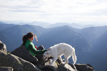 Wanderer, der seinen Hund streichelt, während er sich auf einem Felsen vor einer Bergkette ausruht - CAVF10555