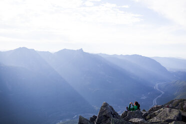 High angle view of woman photographing while sitting on rocks against mountain range - CAVF10557