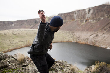 Side view of hiker throwing stone in lake while standing on cliff - CAVF10587