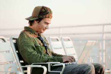Mann, der einen Laptop benutzt, während er auf einem Liegestuhl auf einem Schiff sitzt - CAVF10603