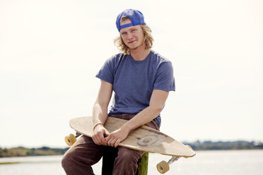 Portrait of man with longboard sitting on wooden post against clear sky - CAVF10651