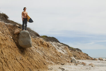 Mann mit Surfbrett auf einem felsigen Berg gegen den Himmel stehend - CAVF10655