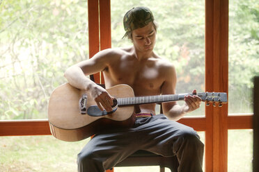 Mann spielt Akustikgitarre, während er zu Hause am Fenster sitzt - CAVF10682