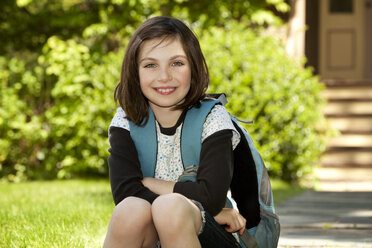 Portrait of happy girl with backpack sitting on footpath - CAVF10714