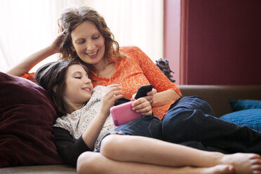 Mutter und Tochter benutzen ein Mobiltelefon, während sie auf dem Sofa sitzen - CAVF10716
