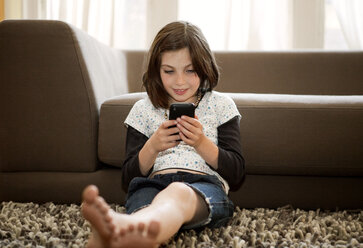 Happy girl using mobile phone while sitting against sofa - CAVF10717