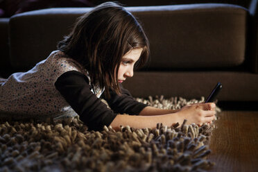 Mädchen benutzt Mobiltelefon, während sie zu Hause auf dem Teppich liegt - CAVF10719
