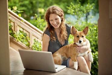 Woman using laptop computer while sitting with dog on porch - CAVF10734