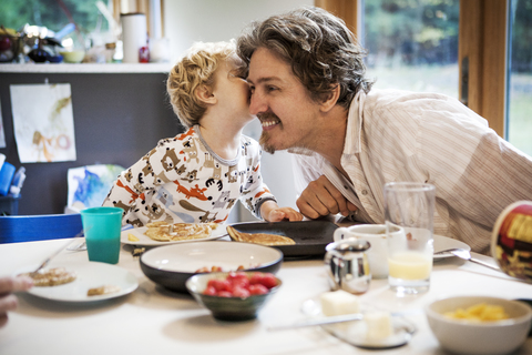 Junge küsst Vater am Esstisch im Haus, lizenzfreies Stockfoto