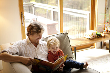 Vater liest mit seinem Sohn ein Buch, während er zu Hause auf dem Sofa sitzt - CAVF10893