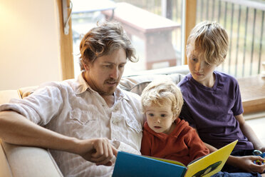 Family reading book while sitting on sofa at home - CAVF10895