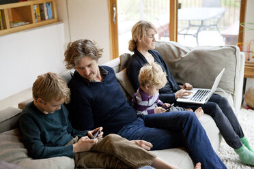 Familie benutzt Laptop und Mobiltelefon, während sie zu Hause auf dem Sofa sitzt - CAVF10930
