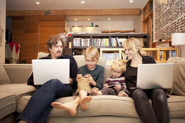 Siblings using mobile phones while sitting with parents on sofa at home - CAVF10932