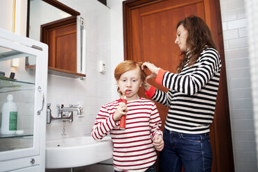 Junge beim Zähneputzen, während die Mutter im Badezimmer die Haare kämmt - CAVF10954