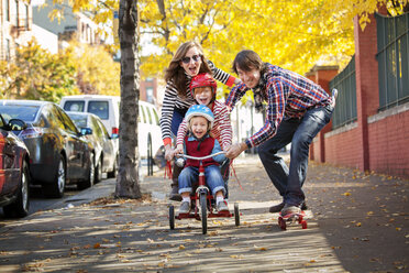 Happy family enjoying skateboard and tricycle on footpath - CAVF10975