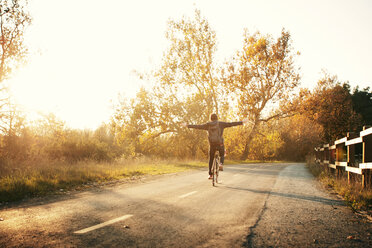 Rückansicht eines Mannes mit ausgestreckten Armen, der bei Sonnenuntergang auf der Straße Fahrrad fährt - CAVF11232