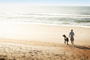 Rückansicht eines Mannes mit Hund, der am Strand spazieren geht - CAVF11247