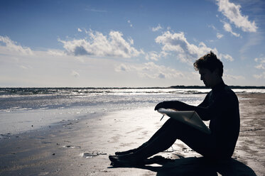 Side view of surfer with surfboard sitting on shore at beach during sunset - CAVF11268