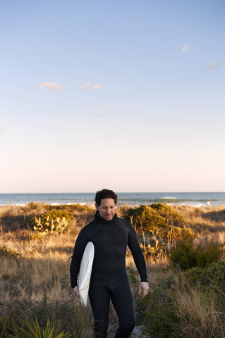 Surfer, der ein Surfbrett trägt, während er auf einem grasbewachsenen Feld gegen den Himmel läuft, lizenzfreies Stockfoto