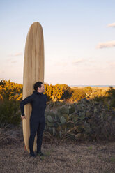 Surfer schaut weg, während er ein Surfbrett auf einer Wiese gegen den Himmel hält - CAVF11279
