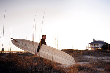 Seitenansicht eines Surfers, der ein Surfbrett trägt, während er auf einer Wiese vor einem klaren Himmel steht - CAVF11283