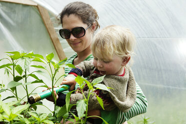 Mutter trägt ihren Sohn beim Gießen von Pflanzen mit dem Gartenschlauch im Gewächshaus - CAVF11377