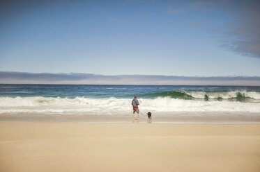 Rear view of man standing with dog on shore against sky - CAVF11436