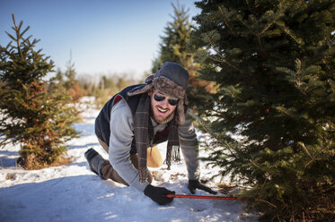 Glücklicher Mann beim Schneiden des Weihnachtsbaums auf einem schneebedeckten Feld - CAVF11443