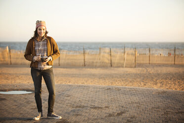 Portrait of woman holding digital camera while standing on sidewalk of beach - CAVF11635