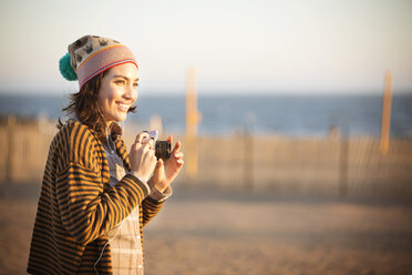 Fröhliche Frau mit Digitalkamera am Strand stehend - CAVF11636