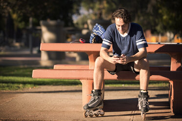 Roller hockey player using mobile phone while sitting on park bench - CAVF11647