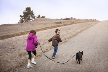 Geschwisterkind mit Hund läuft auf der Straße am Feld gegen den klaren Himmel - CAVF11683