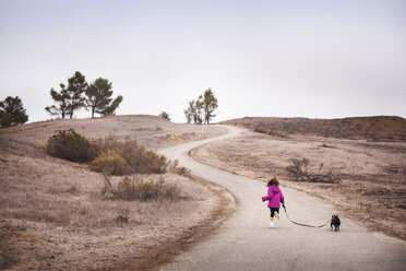 Rear view of girl with dog running on road by field against clear sky - CAVF11684