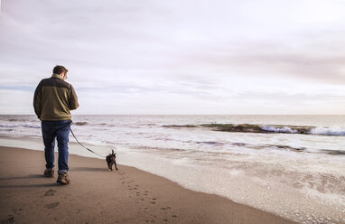 Rückansicht eines Mannes mit Hund, der am Ufer gegen einen bewölkten Himmel spazieren geht - CAVF11689