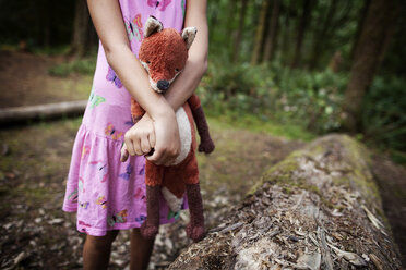 Midsection of girl holding stuffed toy by fallen tree in forest - CAVF11751