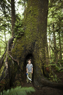 Porträt eines Jungen, der ein Spielzeugschwert hält, während er gegen einen Baumstamm im Wald steht - CAVF11765