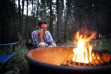 Mann benutzt Mobiltelefon, während er am Lagerfeuer im Wald sitzt - CAVF11785