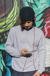 Young man in front of graffity wall hearing music with headphones while using cell phone - AFVF00310