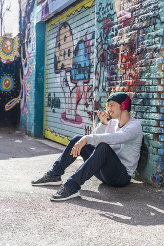 Young man with headphones sitting in front of graffiti wall using cell phone stock photo