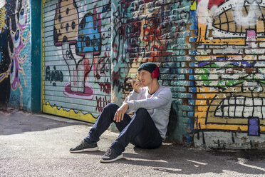 Junger Mann mit Kopfhörern, der vor einer Graffiti-Wand sitzt und ein Mobiltelefon benutzt - AFVF00312